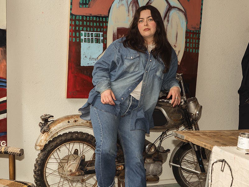 Maddie posing in front of a vintage motorcycle and painting wearing a Wrangler Denim button up and Levi 501 jeans.