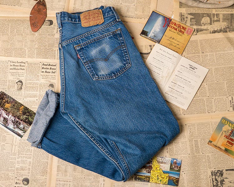 Levi’s blue denim jeans folded to show the back pocket, placed on vintage newspaper pages with postcards, a booklet, and a pair of socks nearby.