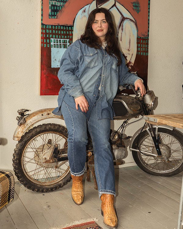 Maddie wearing a Wrangler denim shirt, Levi 501 jeans, and Dan Post brown boots leaning on a vintage motorcycle indoors, with a large painting and wooden table in the background.
