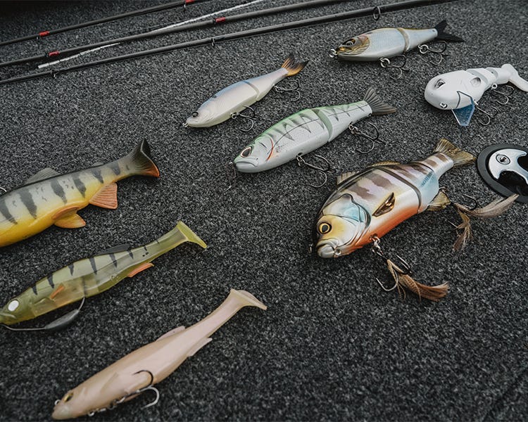 8 different types of swimbaits laying on the deck of a bass boat, soft swimbaits, glide baits, wake baits, from brands like megabass, huddleston, jackall, arashi, 6th sense fishing, strike king, and throwback baits.