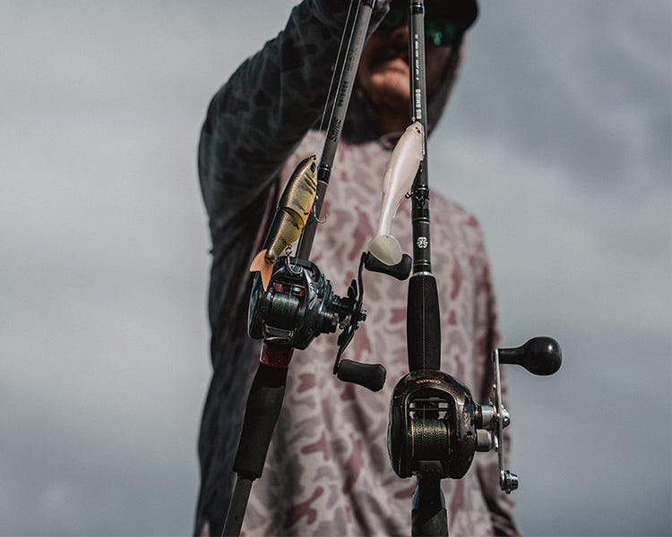 chris standing on the deck of his boat, holding up both of his swimbait rods, the swate big amigo, and the swate brigade black label, with a megabass magdraft and throwback baits tied on
