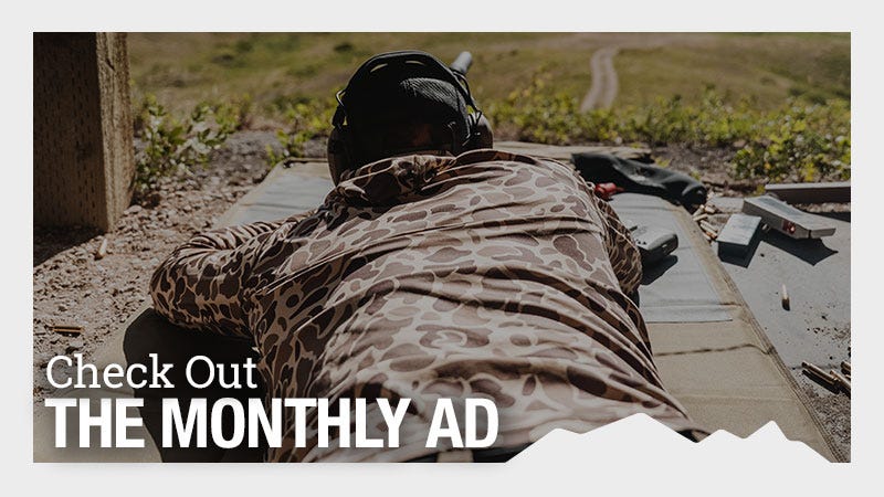 A person wearing camouflage clothing and noise-canceling ear protection lies prone on a shooting mat outdoors, aiming at a distant target. Overlay text reads "Check Out THE MONTHLY AD" in large, bold letters.