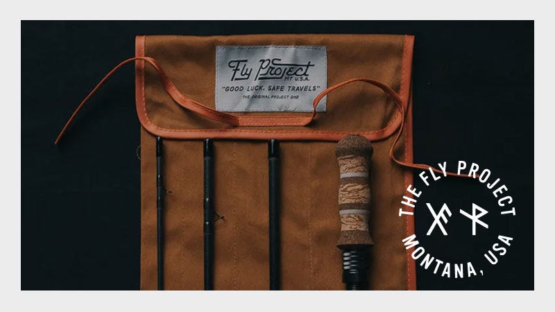 A brown canvas case holds disassembled black fishing rod pieces with a cork handle, labeled "Fly Project" on a patch. The logo for The Fly Project, Montana, USA, appears in the bottom right corner.