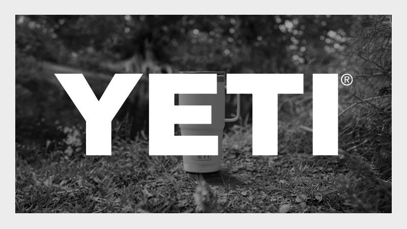 A large white "YETI" logo overlays a black-and-white outdoor scene with a YETI mug placed on grassy ground near a pond and surrounded by foliage.