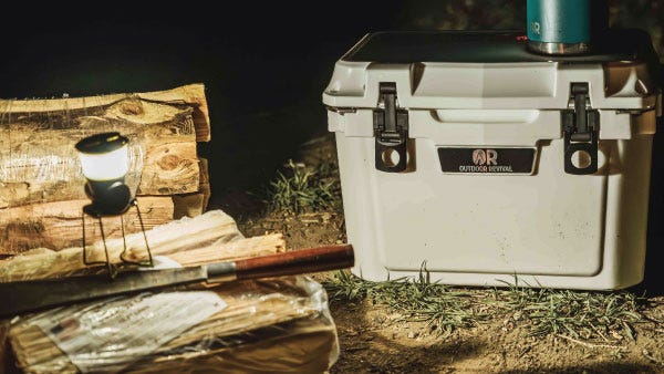 An Outdoor Revival cooler sits on the ground basking in the firelight next to a camp lantern