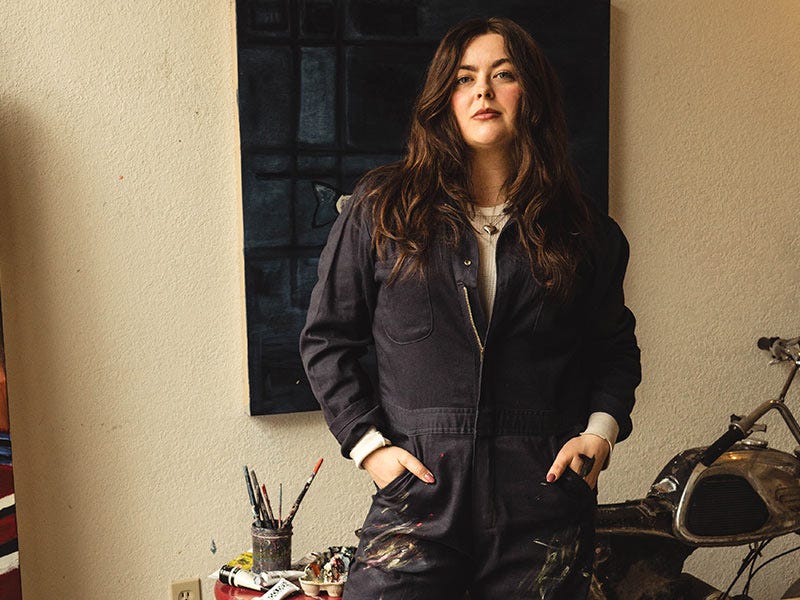 Maddie wearing a Berne coveralls over a light waffle shirt, standing indoors in front of a textured wall and a dark window.
