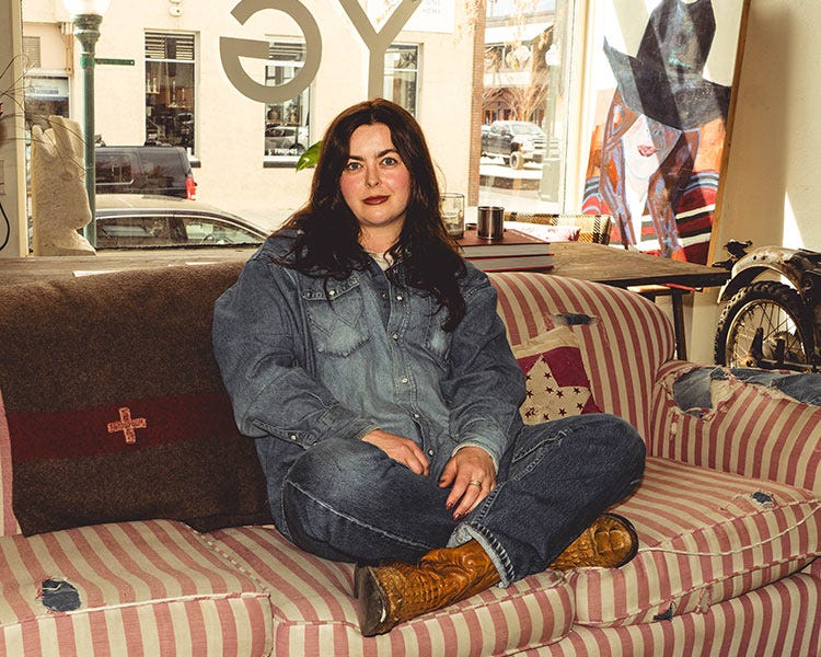Maddie in a Wrangler shirt, Levi jeans, and Dan Post boots sitting cross-legged on a striped couch in a shop, with large windows, artwork, and a motorcycle wheel in the background.