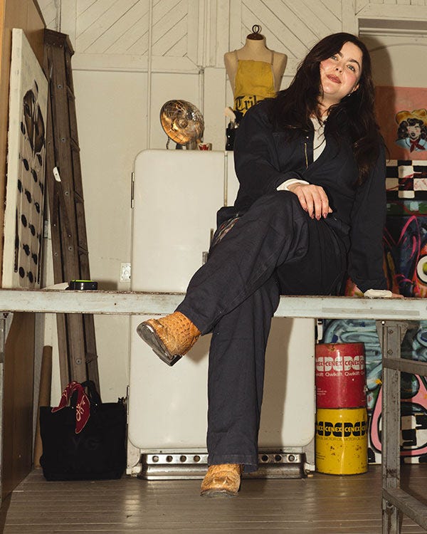 Maddie wearing dark Berne work coveralls and Dan Post boots sitting on a workbench in a studio, with art supplies, a mannequin, and vintage containers in the background.
