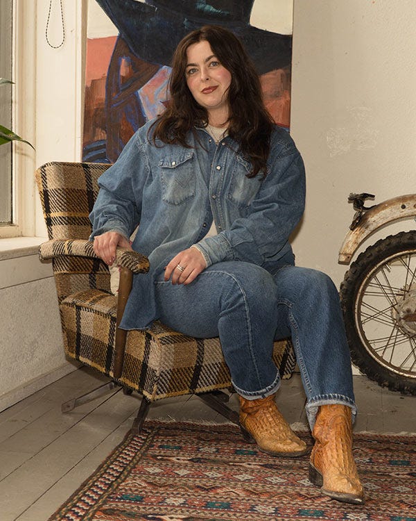 Maddie in a Wrangler denim shirt, Levi jeans, and Dan Post boots seated in a patterned chair, with a painted wall, window, and motorcycle wheel in the background.