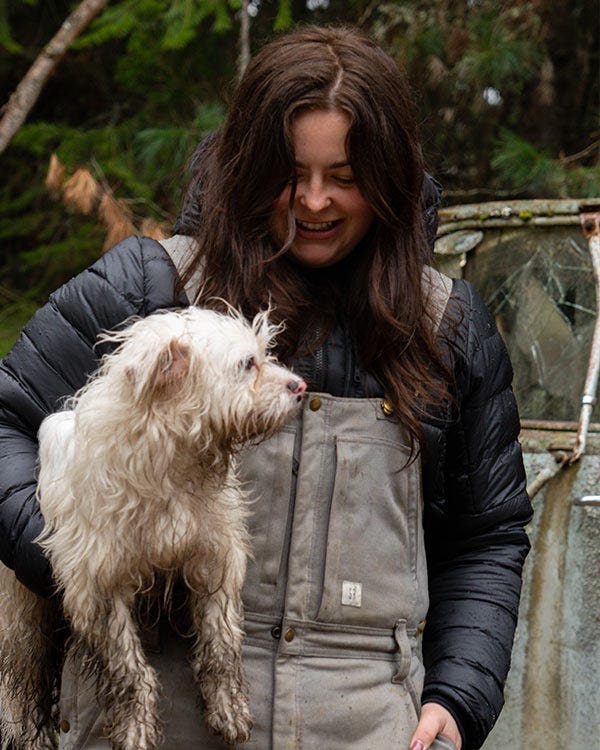 Maddie wearing Smith + Rogue bib overalls and a dark jacket holding a small dog outdoors near a vehicle and trees.