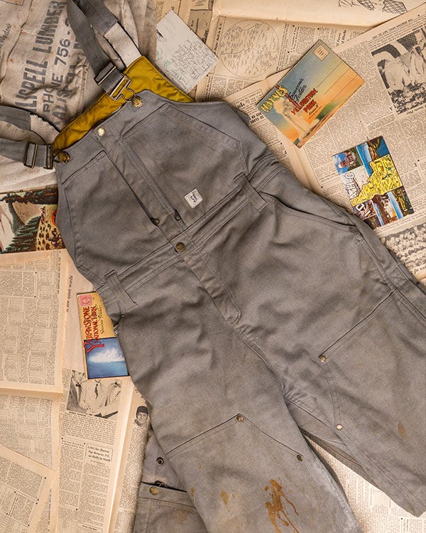 Smith + Rogue gray bib overalls with yellow lining laid on vintage newspaper pages with postcards and a canvas bag.