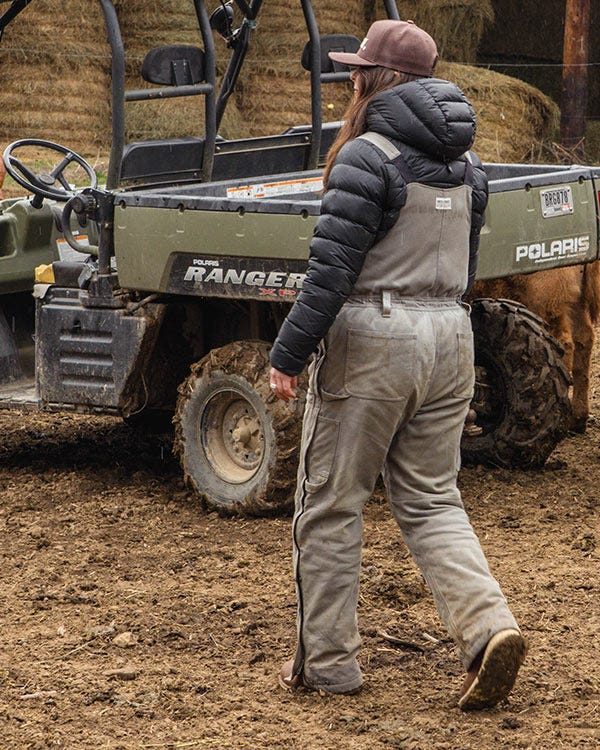Maddie wearing Smith + Rogue bib overalls, a dark jacket, and a cap walking near a Polaris Ranger utility vehicle in a muddy outdoor setting.