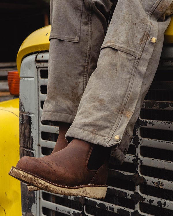 Close-up of brown Danner boots and mud-stained Smith + Rogue overalls resting against the front grille of a yellow vehicle.