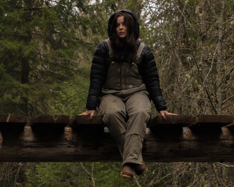 Maddie wearing a dark hooded Smith + Rogue 777 puffy and light-colored bib overalls sitting on a wooden bridge in a forest setting.