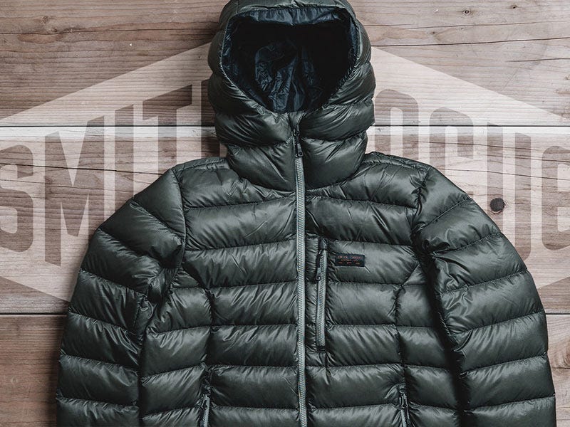 Front view of black Smith + Rogue 777 Front Range puffer jacket with insulated quilted design, hood, front zipper closure, and zip chest pocket displayed on a wood backdrop.