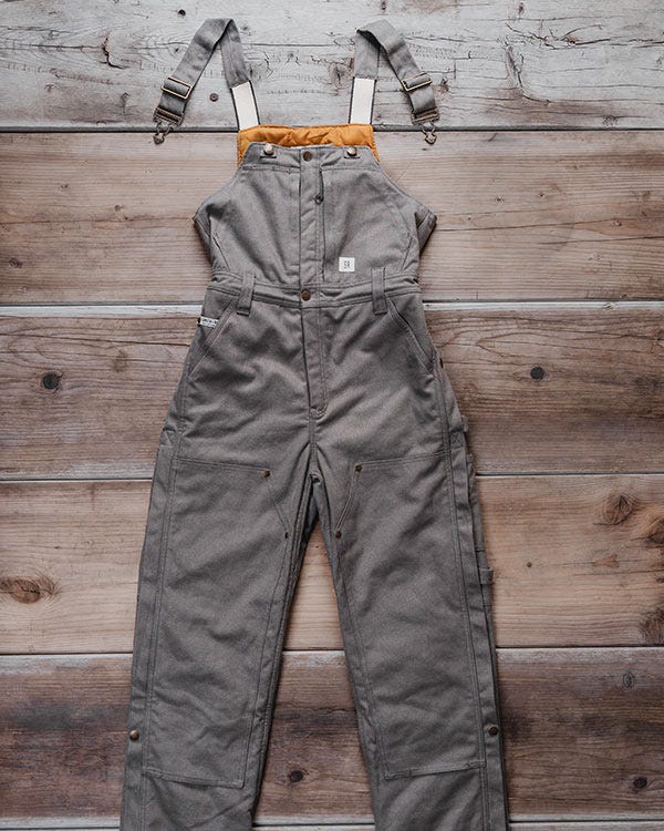 Smith + Rogue Original canvas bib overalls in gray with adjustable straps, shown laid flat on a wooden surface.