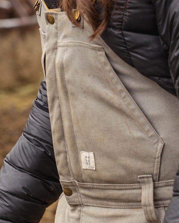 Close-up of Smith + Rogue Original canvas bib overalls showing chest pocket, buttons, and logo patch over a dark jacket.