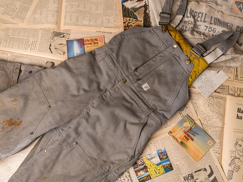 Gray Smith and Rogue canvas work overalls with metal hardware are laid flat across vintage newspapers, postcards, and printed materials, showing multiple pockets, adjustable straps, and worn paint marks on the fabric.