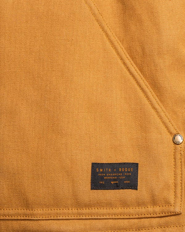 Close-up of Smith + Rogue original canvas material showing tan heavyweight canvas fabric with reinforced stitching and woven Smith + Rogue logo patch near the hem.