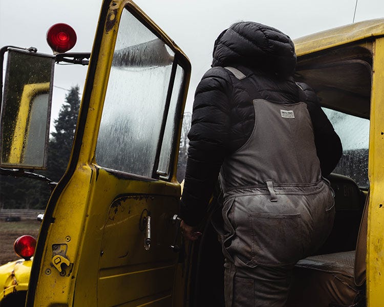 Person wearing gray insulated Smith + Rogue canvas bib overalls and a black hooded puffer jacket while climbing into a yellow utility vehicle on a rainy day.