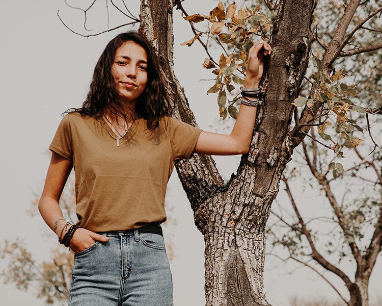 Person wearing a tan Smith + Rogue Pima cotton t-shirt and jeans while standing beside a tree outdoors in a natural setting.