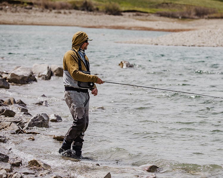 Person fly fishing along a rocky shoreline while wearing Smith + Rogue Skoltec hoodie and chest waders beside a moving lake.