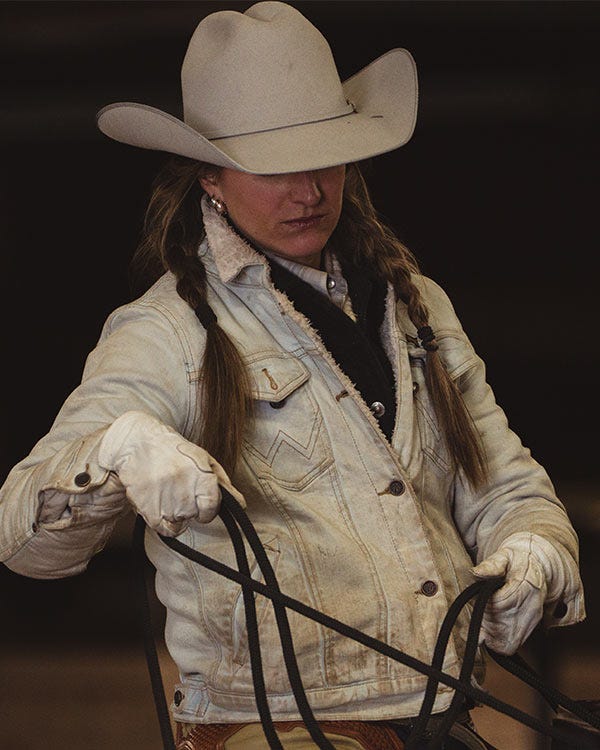 Woman wearing a light-colored cowboy hat and denim jacket holds leather reins while looking down, photographed against a dark background.