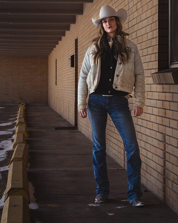 Woman wearing a cowboy hat, denim jacket, and jeans stands in a covered outdoor walkway with brick walls and patches of snow on the ground.