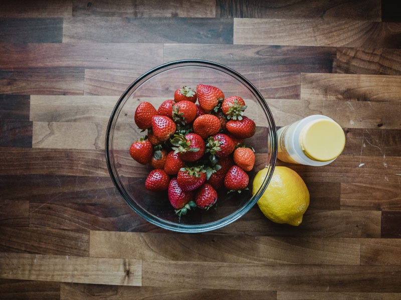 Fresh Florida strawberries in a clear glass bowl sit next to a lemon and a jar of Smoot honey on a butcher block