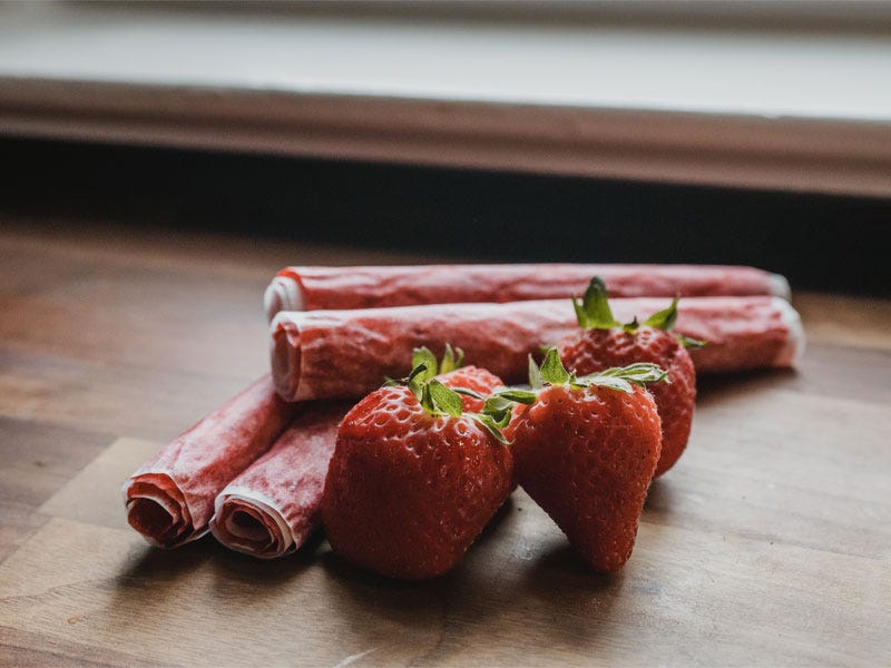 Finished and rolled fruit leathers sitting next to fresh strawberries in bright window light