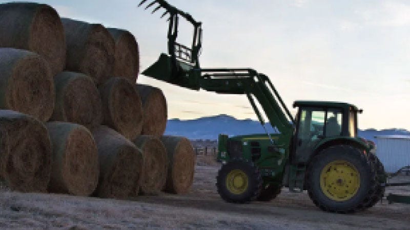 A John Deer tractor stacks hay bails with during sunset with mountains in the background