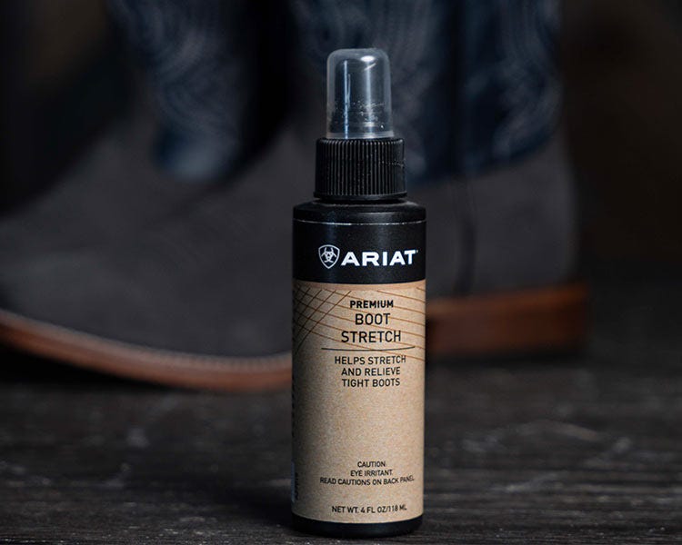 Bottle of Ariat Premium Boot Stretch spray standing upright on a dark surface, with a pair of boots blurred in the background.