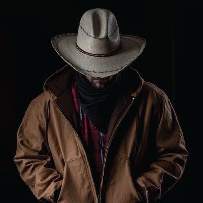 A dramatically lit man wearing a brown fabric work jacket and Montana style straw cowboy hat