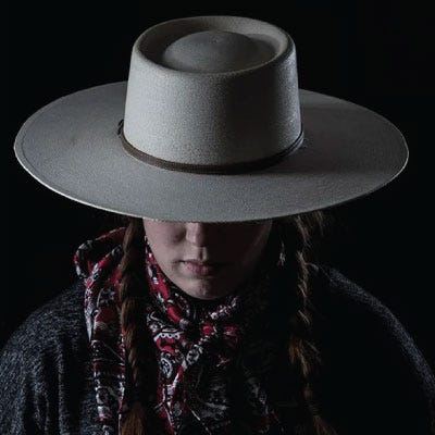 A dramatically photo of a woman with braids wearing a gambler style straw cowboy hat 