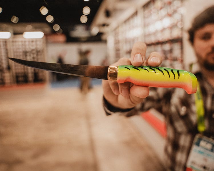 Rapala Firetiger Filet Knife with classic green and orange handle, stainless steel blade reflecting light on a fillet table with fish nearby. Held in hand at the Rapala booth at the 2025 ICAST show. 