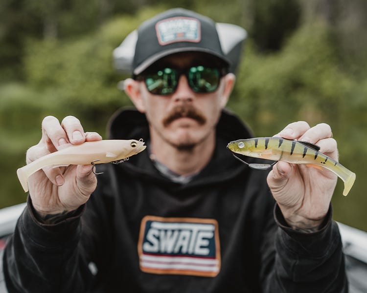chris sitting on the deck of his boat holding up two soft swimbaits, the megabass magdraft and the megabass magdraft freestyle