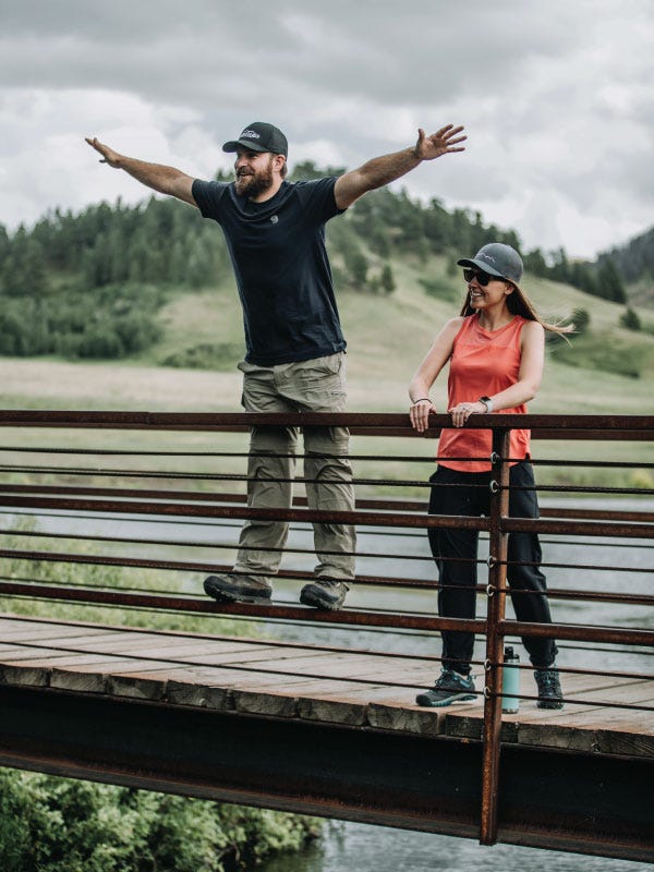 A man and women stand on a bridge during a hike. The man does a "Titanic" pose while the women looks on smiling.
