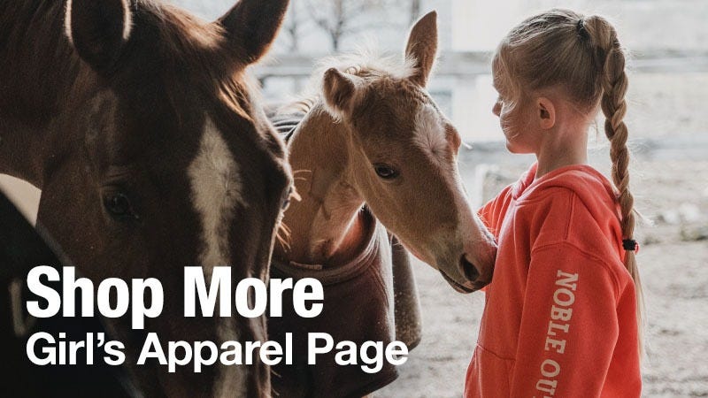 A young girl with a long braid, wearing a red hoodie that says "NOBLE OUT," stands indoors gently touching the face of a light brown foal beside a dark brown horse. Large white text over the image reads "Shop More Girl&rsquo;s Apparel Page."