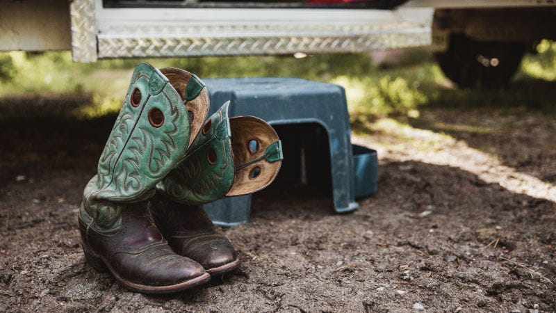 A pair of brown and green round toe cowboy boots sit in the wet dirt next to a trailer step and stool